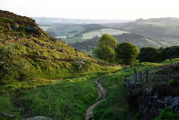 Peak district path This landscape photograph shows a rural nature path winding through green fields and gently sloping hills in the Peak District, United Kingdom, specifically in Derbyshire near Curbar Edge. The image was captured during the evening in late spring, which is indicated by the vibrant green foliage on the trees, the lush grass, and the soft golden light cast over the landscape. In the foreground, dry stone walls and wire fencing are typical features of the Derbyshire countryside, while the background reveals a patchwork of fields and wooded areas leading towards distant hills. The photograph demonstrates the natural beauty of the Peak District, highlighting its scenic rural environment and the characteristic terrain of Curbar Edge.